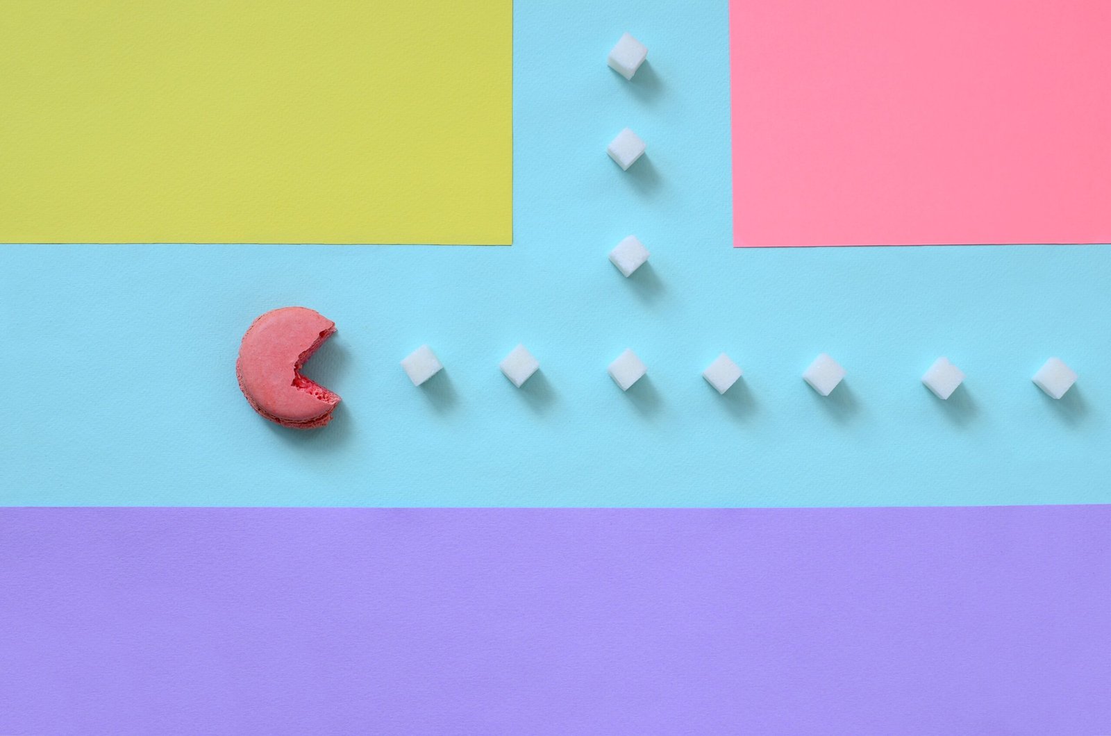 A composition of macaroon with an open mouth that is going to eat sugar cubes on a blue, pink, yellow and violet pastel background. The concept of old video games
